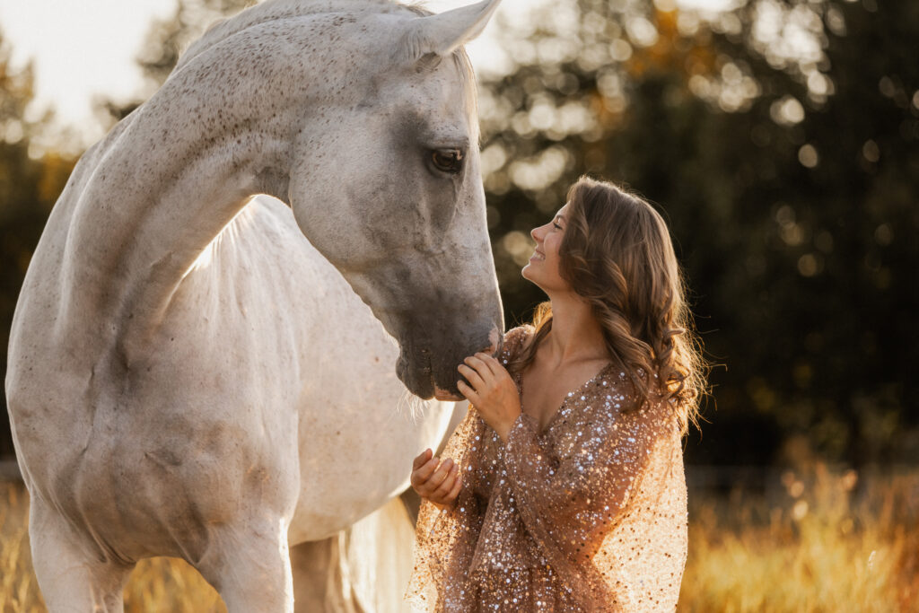 weißer Schimmel und Frau im Glitzerkleid in der Abendsonne